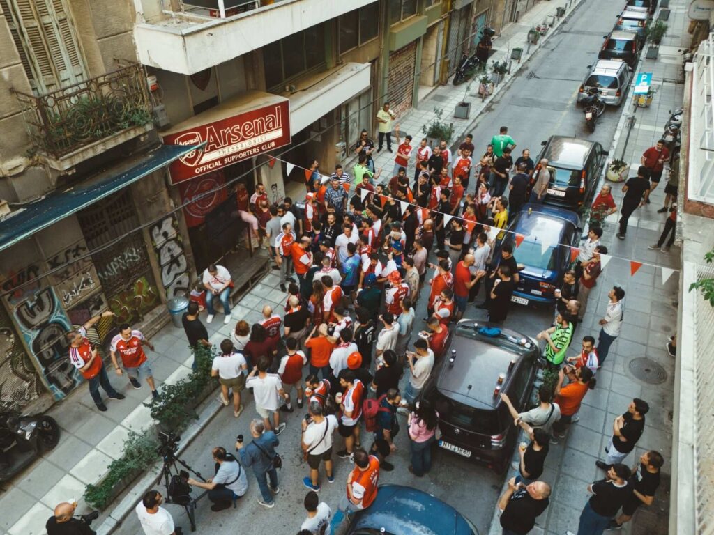 A drone shot of a large crowd of Arsenal supporters gathers outside the Arsenal Thessaloniki Official Supporters Club on a narrow city street.