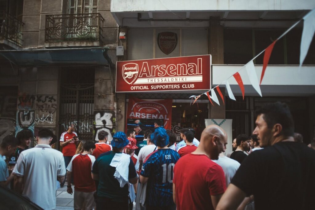 Arsenal supporters gather outside the Arsenal Thessaloniki Official Supporters Club beneath its red sign and red-and-white bunting.